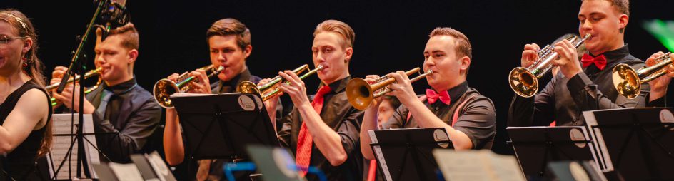 Musiker spielen Trompeten auf der Bühne, umrahmt von Notenständern und Mikrofonen.