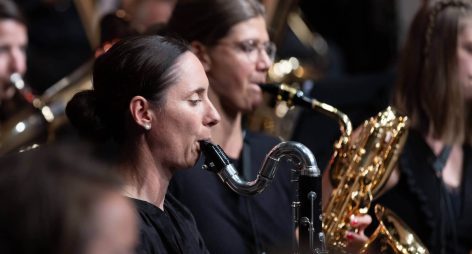 Musikerinnen spielen Blasinstrumente in einem Blasorchester, im Hintergrund weitere Musiker.