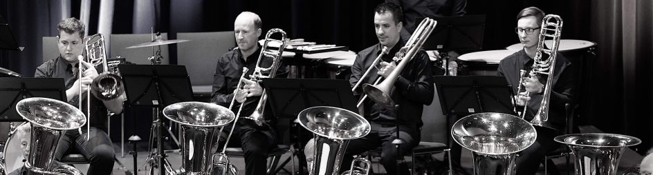 Blasmusikensemble mit mehreren Musikern an Tubas und Posaunen auf der Bühne.