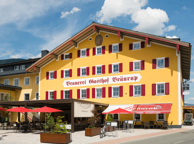 Gelbes Alpenhotel mit rotem Schriftzug, Terrasse, Sonnenschirmen und Bergkulisse