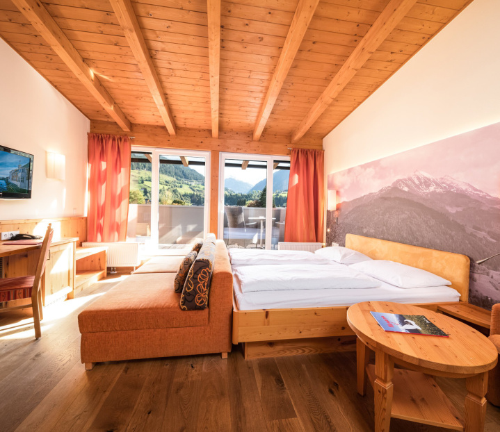 Helles Hotelzimmer mit Holzmöbeln, Doppelbett, Sofa und Balkonblick auf die Alpen