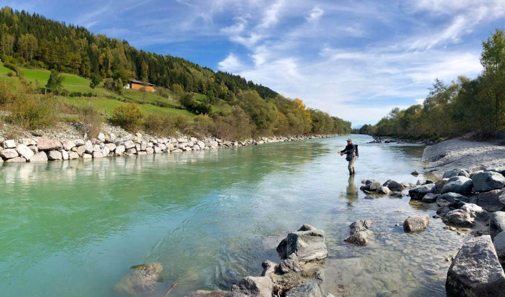 Angler steht am türkisfarbenen Fluss vor Alpenlandschaft und grünem Wald