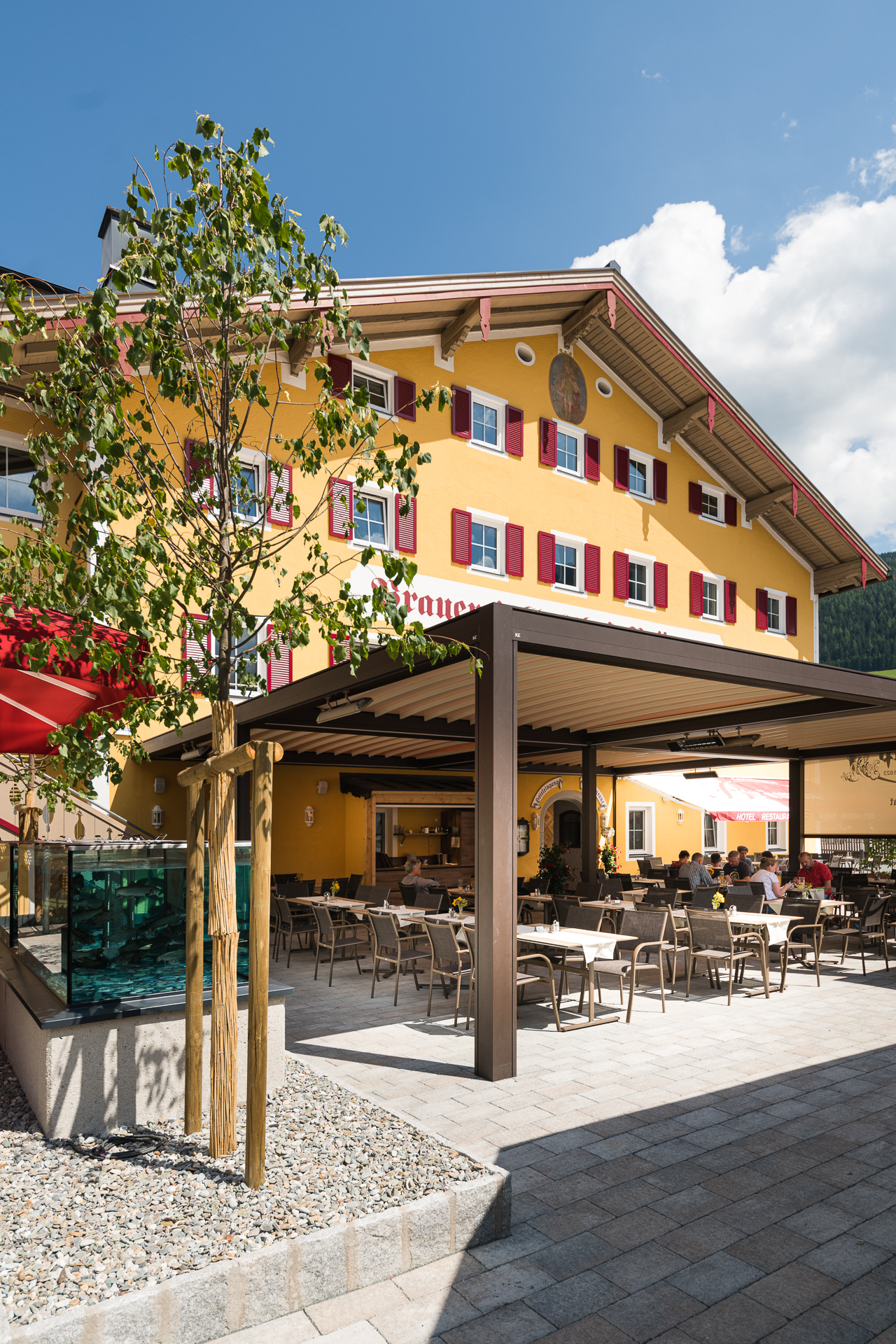 Gelbes Hotelgebäude mit rotem Fensterläden und Terrasse mit Sitzplätzen