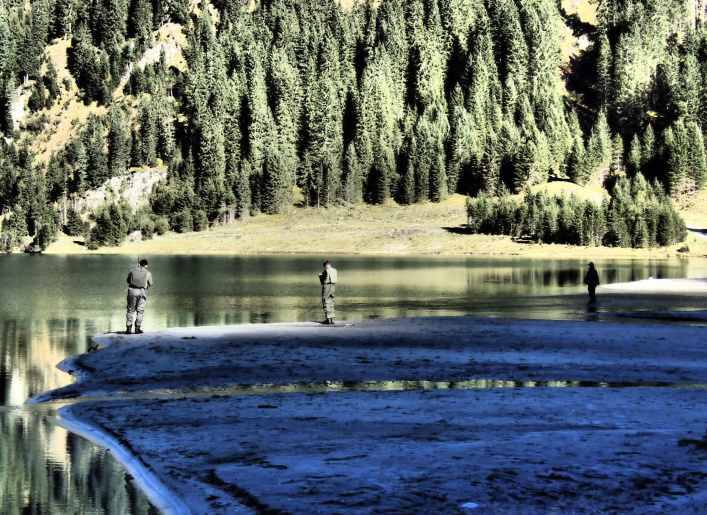 Drei Angler stehen am Ufer eines klaren Bergsees vor dichtem Nadelwald