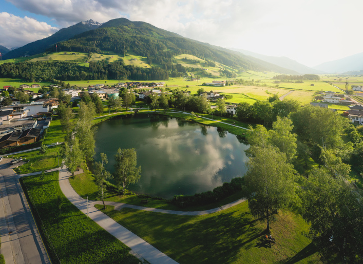 Zierteich mit Wegen und Bäumen in Mittersill vor grünen Wiesen und bewaldeten Bergen
