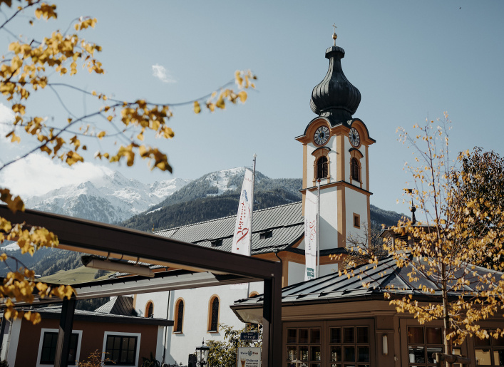 Kirchturm mit Zwiebeldach vor schneebedeckten Bergen und gelben Herbstblättern