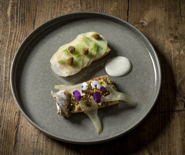 Gourmetgericht mit Fisch, essbaren Blüten und Gemüse auf grauem Teller