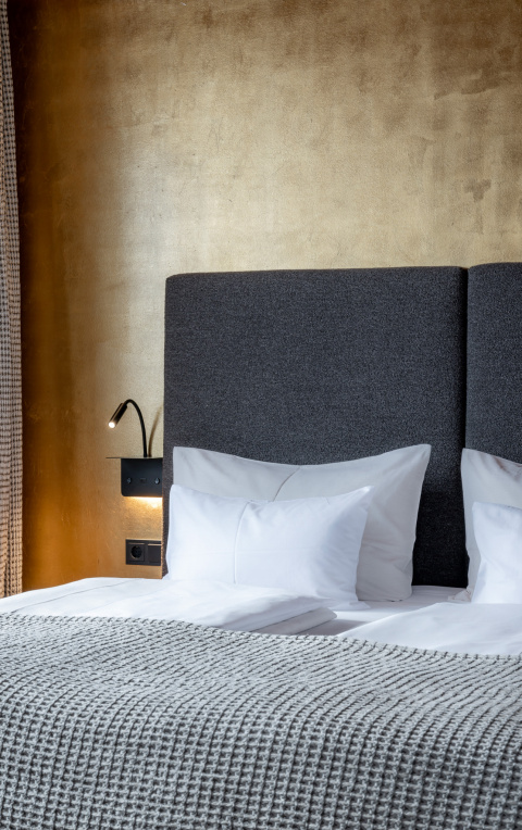 Doppelbett mit weißen Kissen, grauem Kopfteil, goldener Wand und kleiner Leselampe
