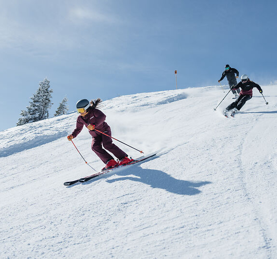 Drei Personen fahren bei Sonne eine schneebedeckte Piste hinab