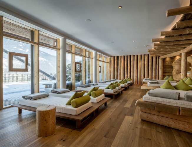 Holzverkleideter Ruheraum mit Liegen, grünen Kissen und großer Fensterfront mit Blick auf verschneite Berge