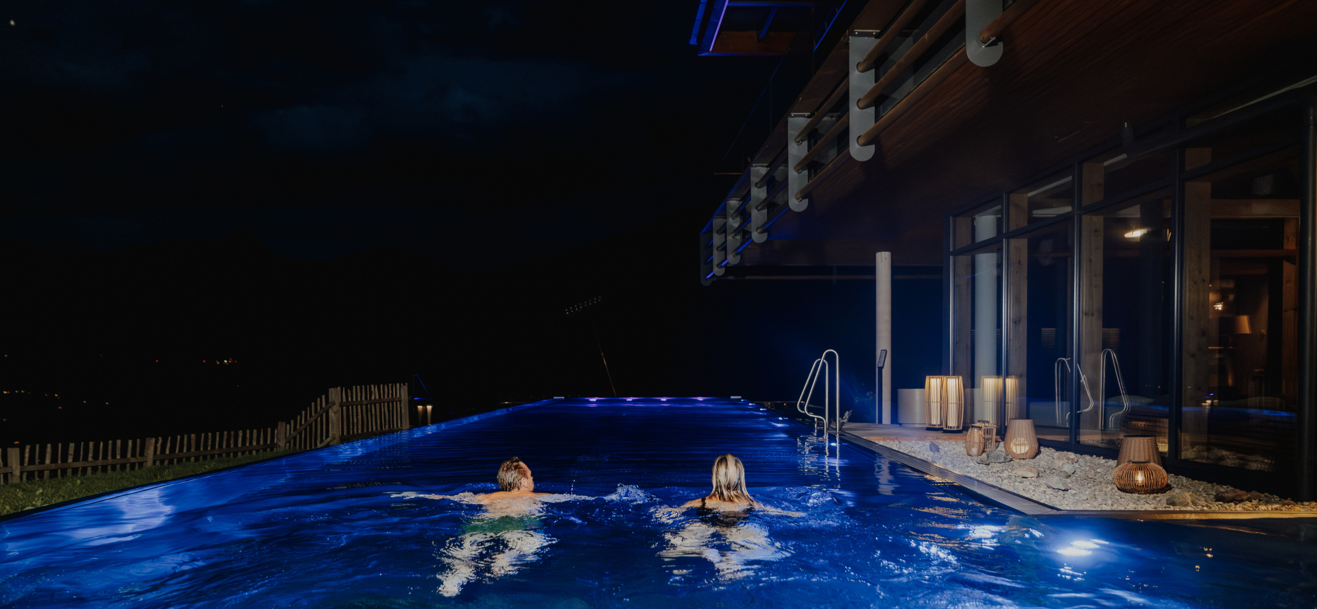 Zwei Personen beim nächtlichen Schwimmen im beleuchteten Infinity-Pool mit Blick ins dunkle Tal