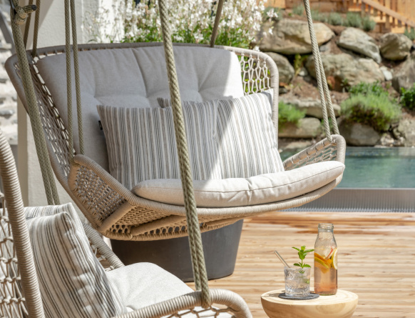 Hängesessel mit Kissen auf sonniger Holzterrasse neben kleinem Tisch mit Getränken und Pool