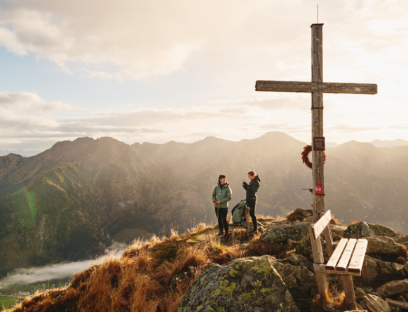 Zwei Wandernde am Gipfelkreuz neben einer Bank mit Blick auf sonnenbeschienene Bergketten
