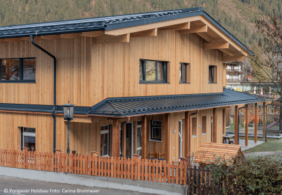 Holzverkleideter Kindergarten mit dunklem Dach und eingezäuntem Garten vor bewaldetem Hang