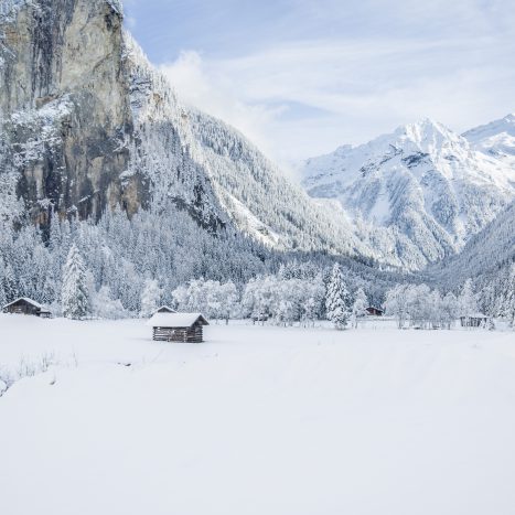 Verschneite Alpenlandschaft mit Holzhütten, Tannenwald und schneebedeckten Bergen