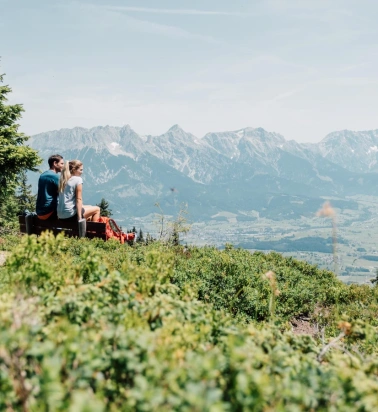 Zwei Wandernde sitzen auf einer Bank mit Blick über ein Tal und eine Bergkette