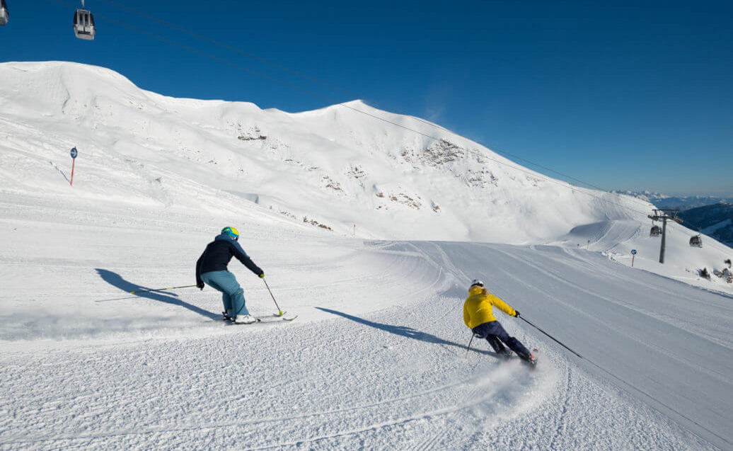 Zwei Skifahrer fahren auf einer sonnigen, präparierten Piste durch verschneite Berglandschaft