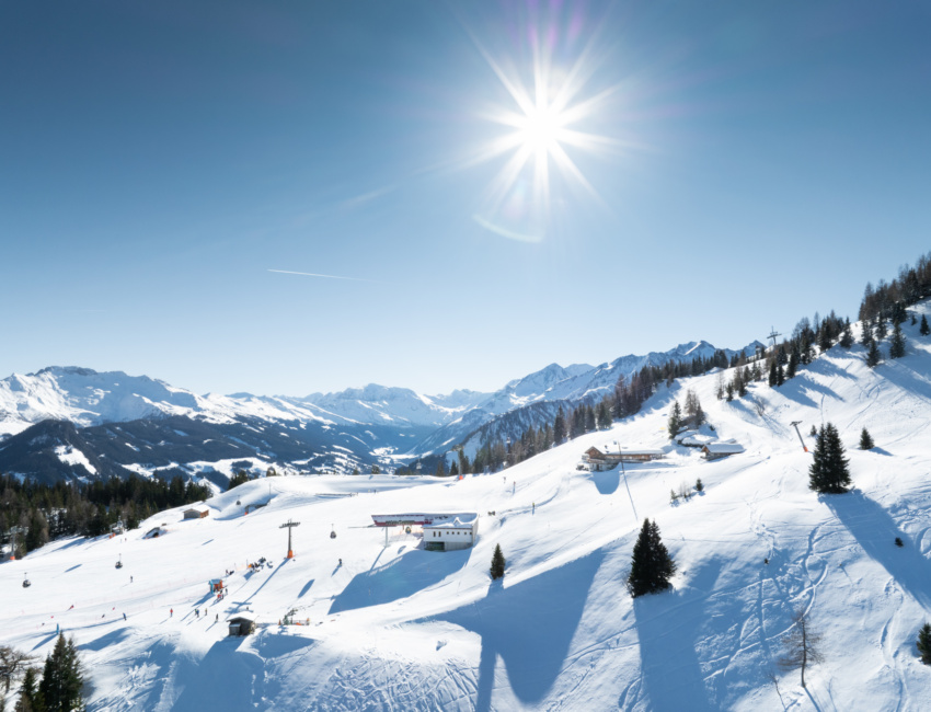 Paesaggio montano innevato con piste da sci, impianti di risalita e singoli sciatori sotto un cielo blu