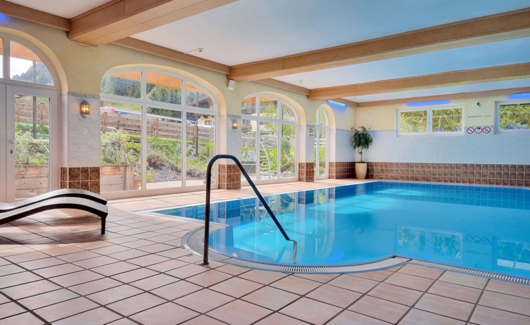 Indoor pool with blue water, lounge chair, wooden ceiling, and large arched windows facing the garden