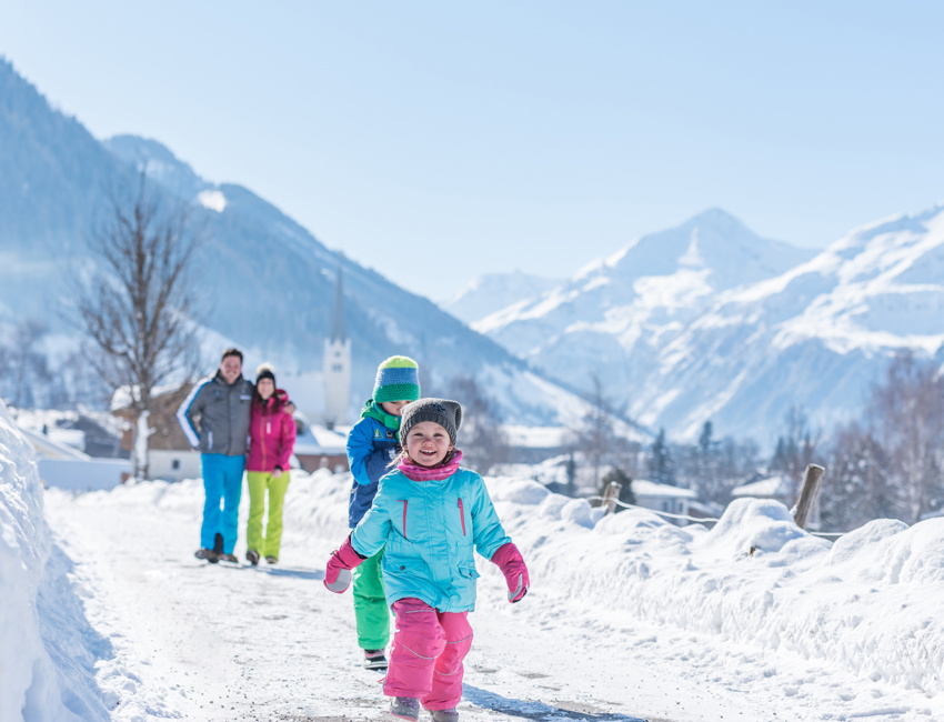 Zwei Kinder und zwei Erwachsene auf einem verschneiten Winterweg vor hohen Bergen