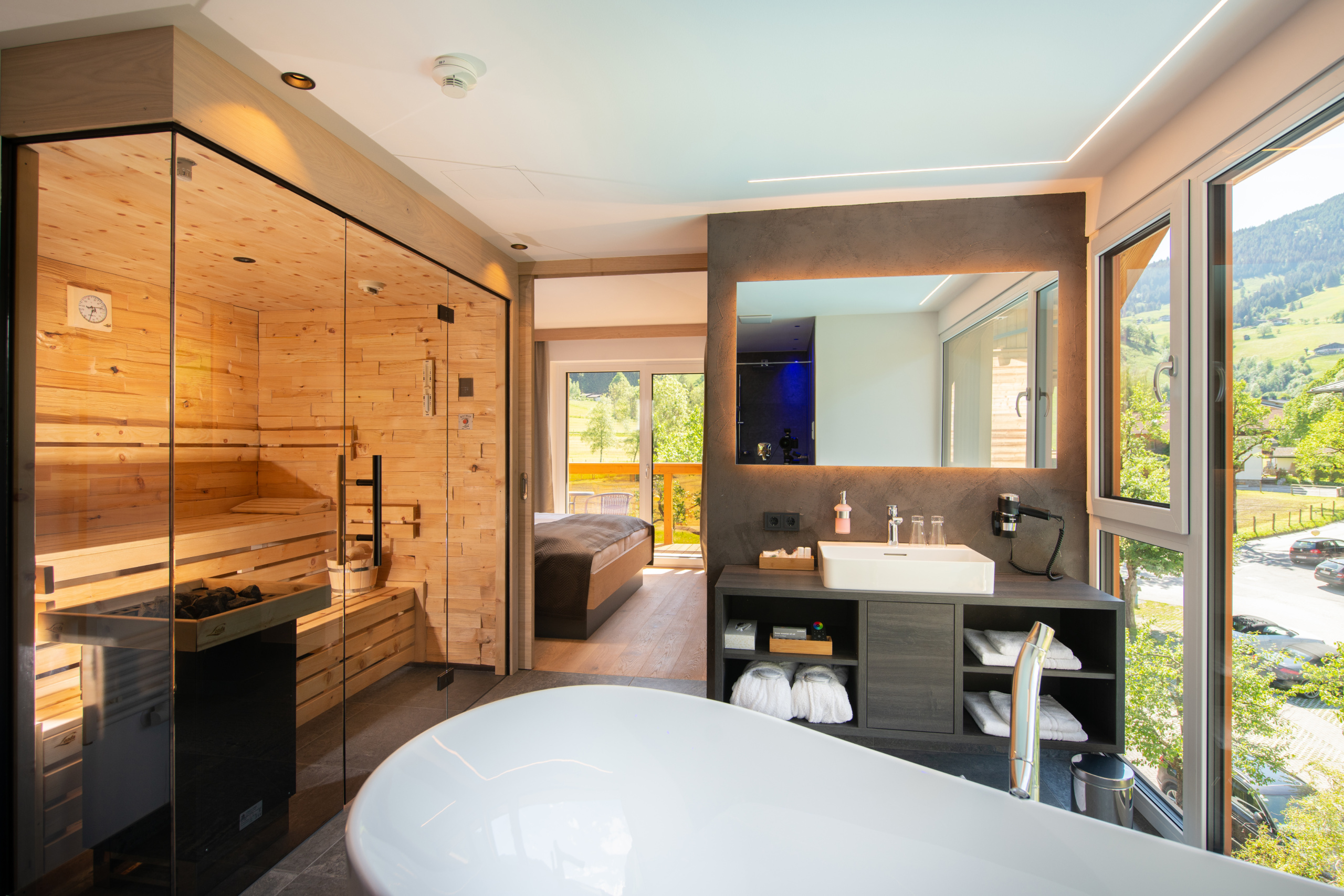 Freistehende Badewanne vor Sauna und Waschtisch in modernem Hotelbad mit Blick ins Grüne