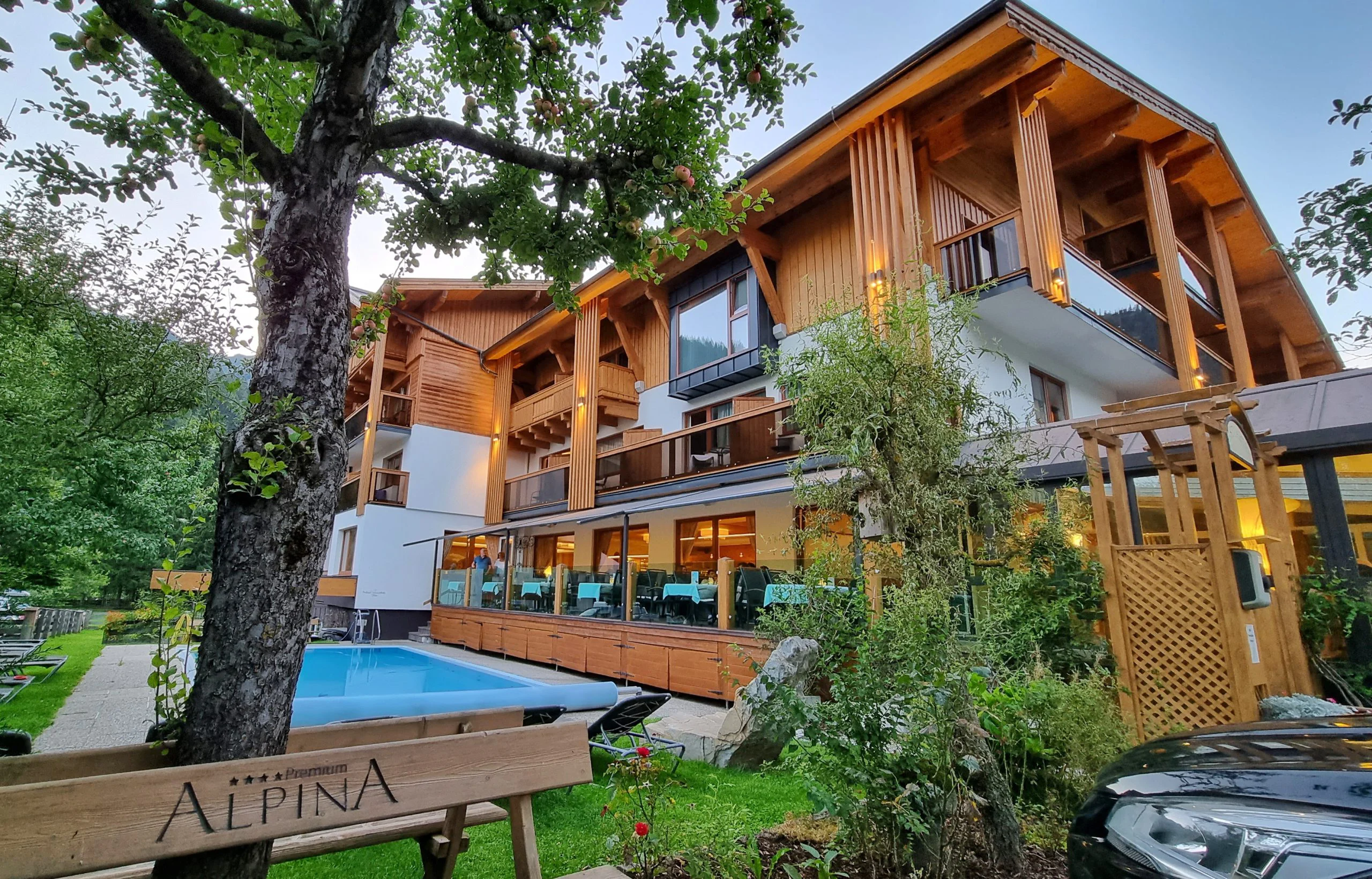 Hotel Alpina s dřevěnou fasádou, balkony, zahradou a venkovním bazénem večer
