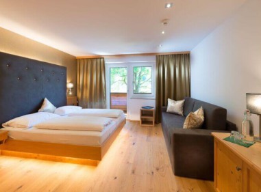 Helles Hotelzimmer mit Doppelbett, dunklem Sofa, Holzmöbeln und Balkonfenster