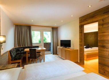 Helles Hotelzimmer mit Doppelbett, Sitzecke, Balkonfenster, Fernseher und offenem Durchgang zu einem weiteren Bett