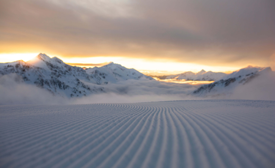 Frisch präparierte Skipiste vor verschneiten Bergen bei Sonnenaufgang