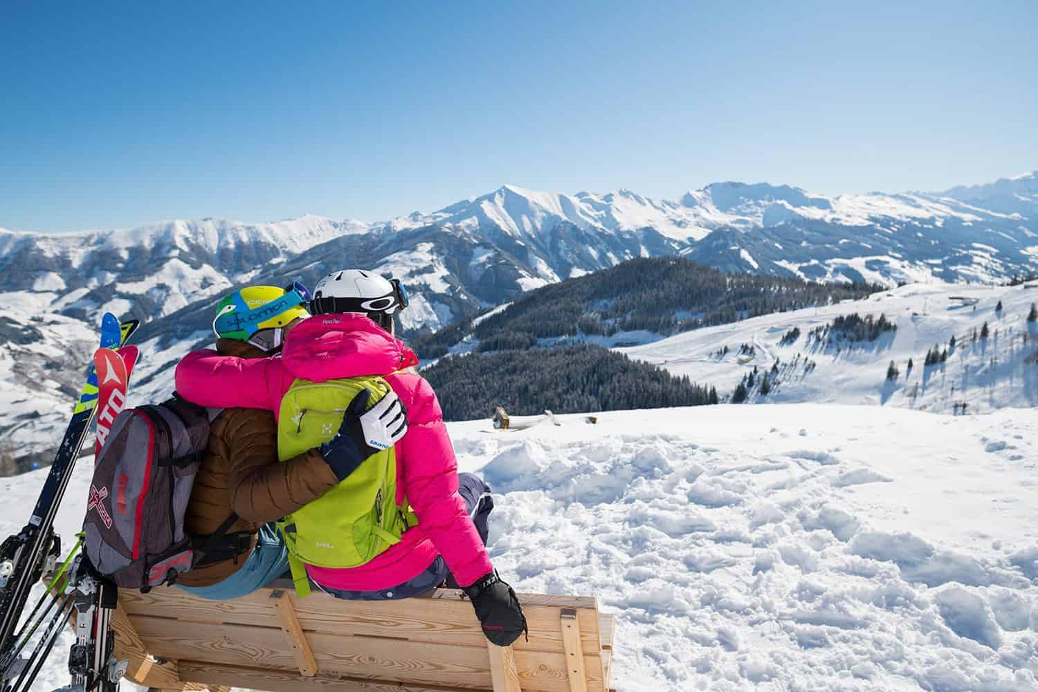 Zwei Skifahrer in bunter Winterkleidung umarmen sich auf einer Bank vor verschneiten Bergen