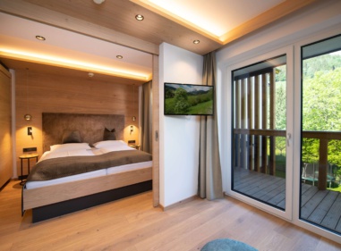 Helles Hotelzimmer mit Doppelbett, Holzverkleidung, Fernseher und Balkon mit Blick ins Grüne