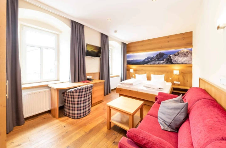 Helles Hotelzimmer mit Doppelbett, Schreibtisch, rotem Sofa und Bergpanorama