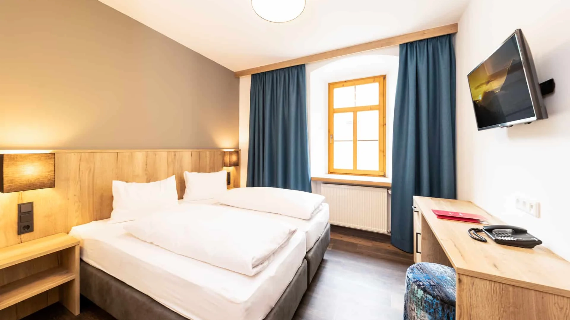 Modernes Hotelzimmer mit Doppelbett, Schreibtisch, TV und Fenster mit blauen Vorhängen