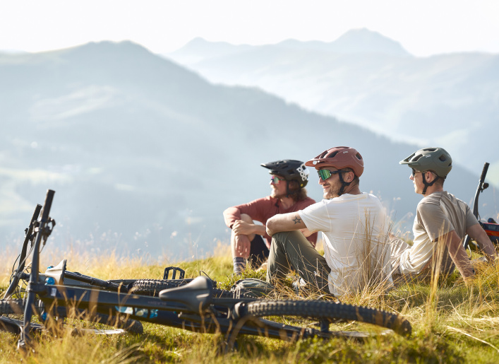 Drei Mountainbiker sitzen auf einer Bergwiese mit Blick ins Tal