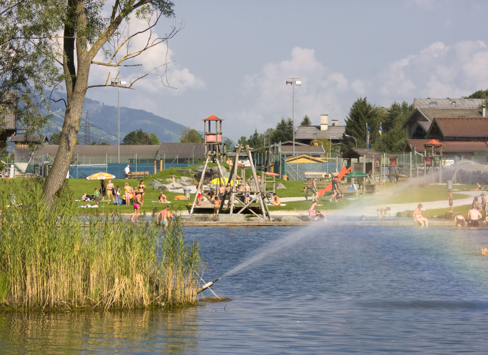 Badesee mit Schilf, Badegästen, Spielplatz und Bergkulisse an einem sonnigen Sommertag