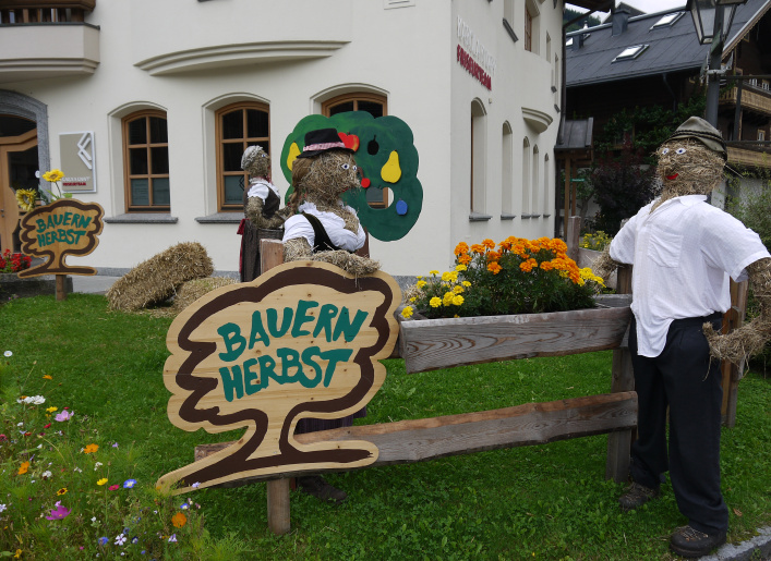 Two straw figures in front of a house with "Bauernherbst" sign and colorful flowers