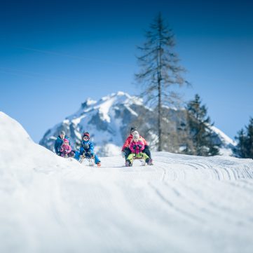 Familie rodelt im Schnee vor verschneitem Bergpanorama und blauem Himmel