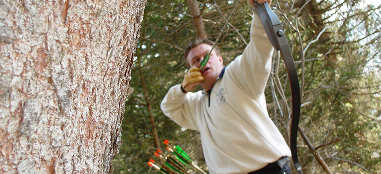 Bogenschütze zielt mit einem Jagdbogen im Wald, umgeben von Pfeilen und Natur.