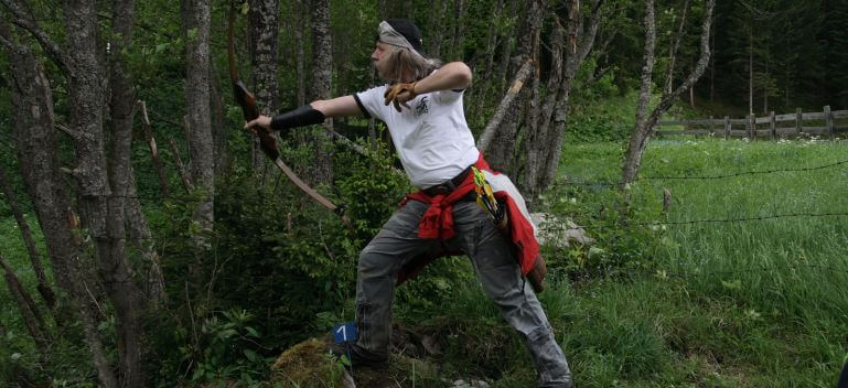 Bogenschütze zielt mit Jagdbogen in der Natur, umgeben von Bäumen und Grün.