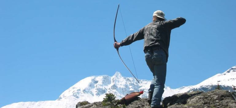 Bogenschütze zielt mit einem Jagdbogen auf eine Zielscheibe vor schneebedeckten Bergen.