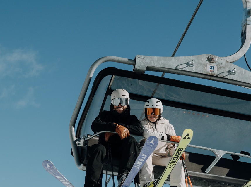 Zwei Skifahrer sitzen mit Skiern und Helmen auf einem Sessellift vor blauem Himmel
