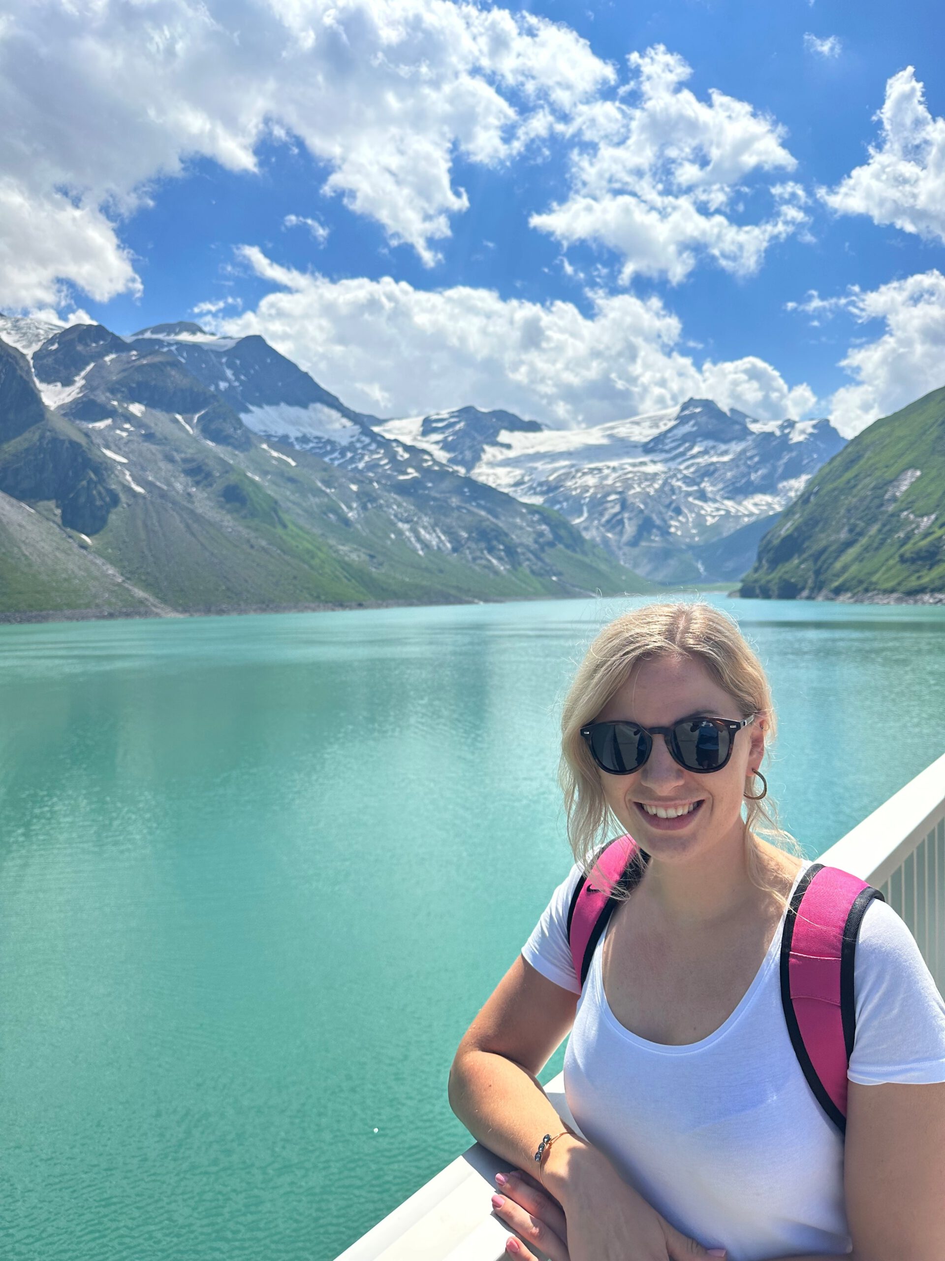 Frau mit Sonnenbrille vor türkisfarbenem Stausee in den Alpen