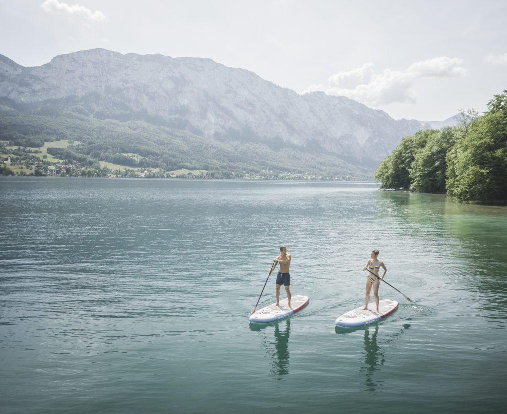 Zwei Personen paddeln auf SUP-Boards am Attersee vor grünen Hügeln im Salzkammergut